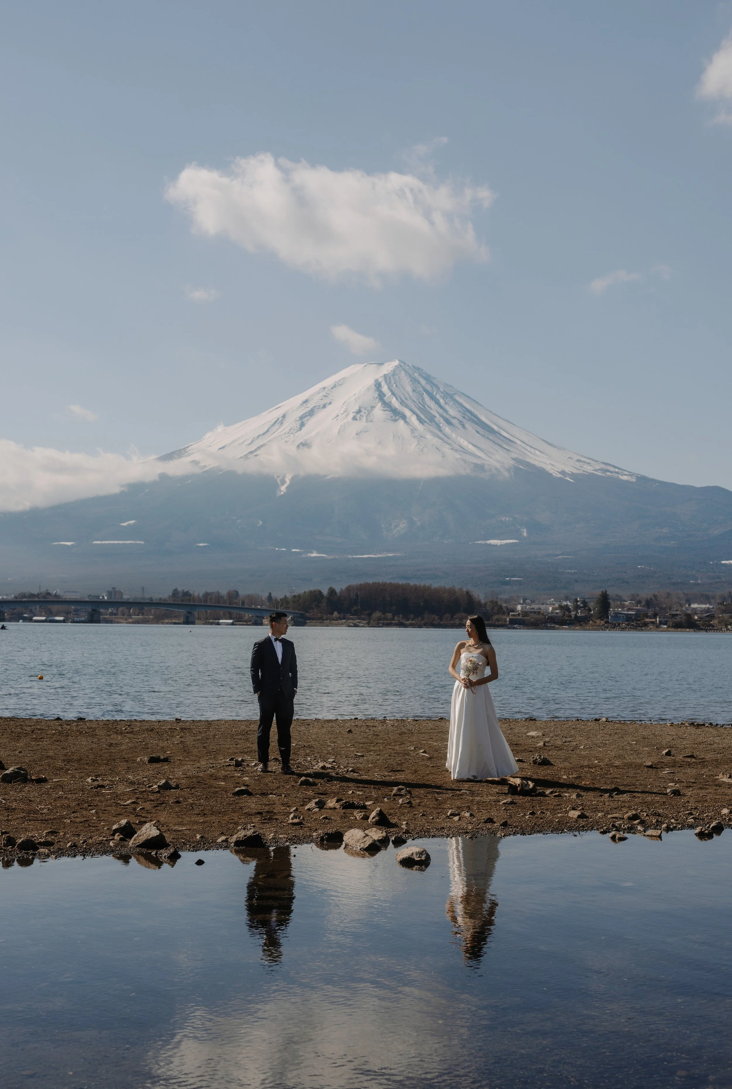 Bride and groom standing apart on a lakeshore with their reflection in the water and snow-capped Mount Fuji behind