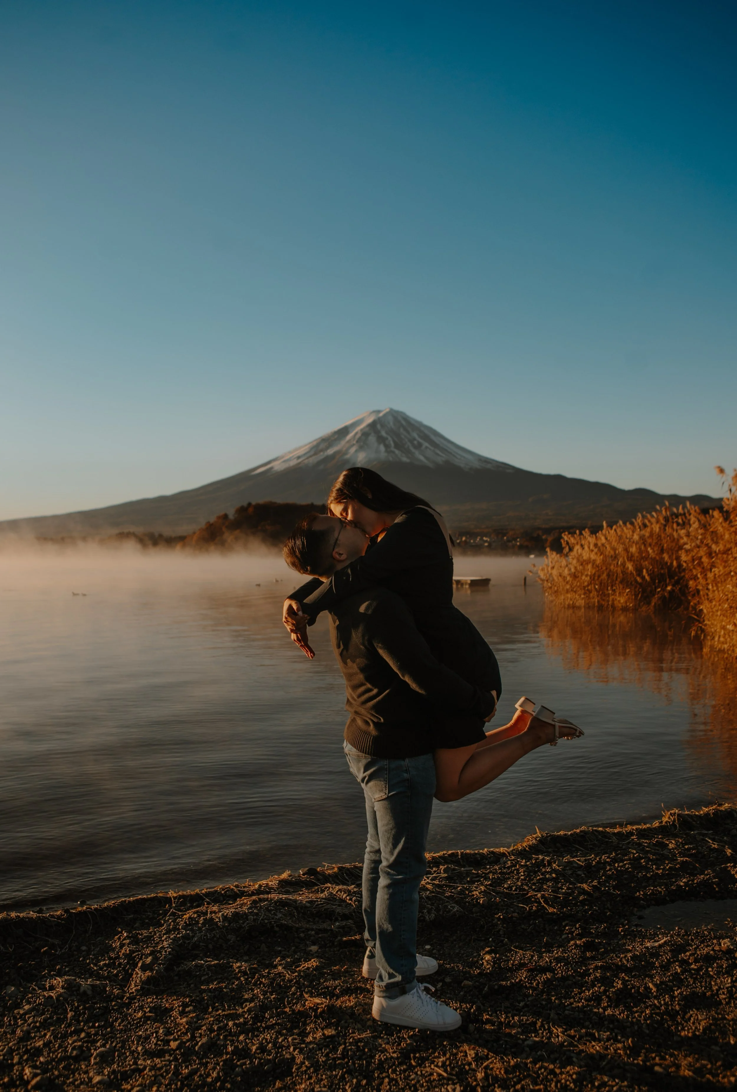 Groom giving bride a playful piggyback ride on the shore of Lake Kawaguchiko with Mount Fuji in the distance