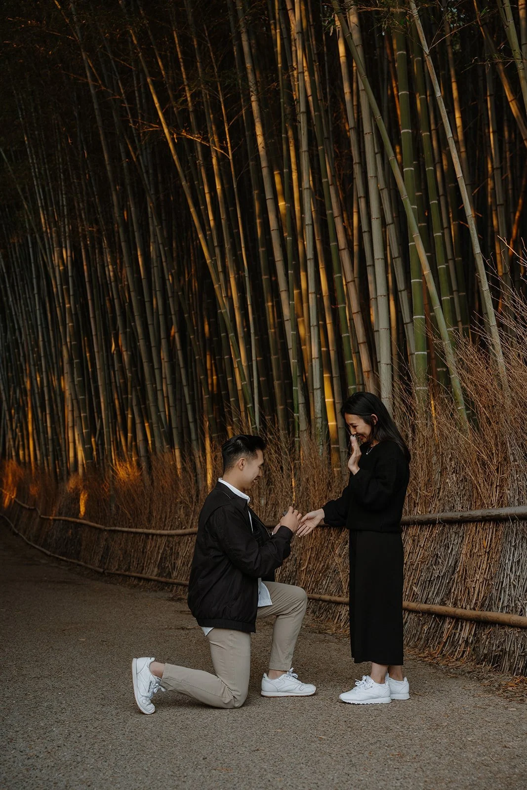 Man kneeling to propose in the Arashiyama Bamboo Grove at golden hour, his partner covering her face in surprise.