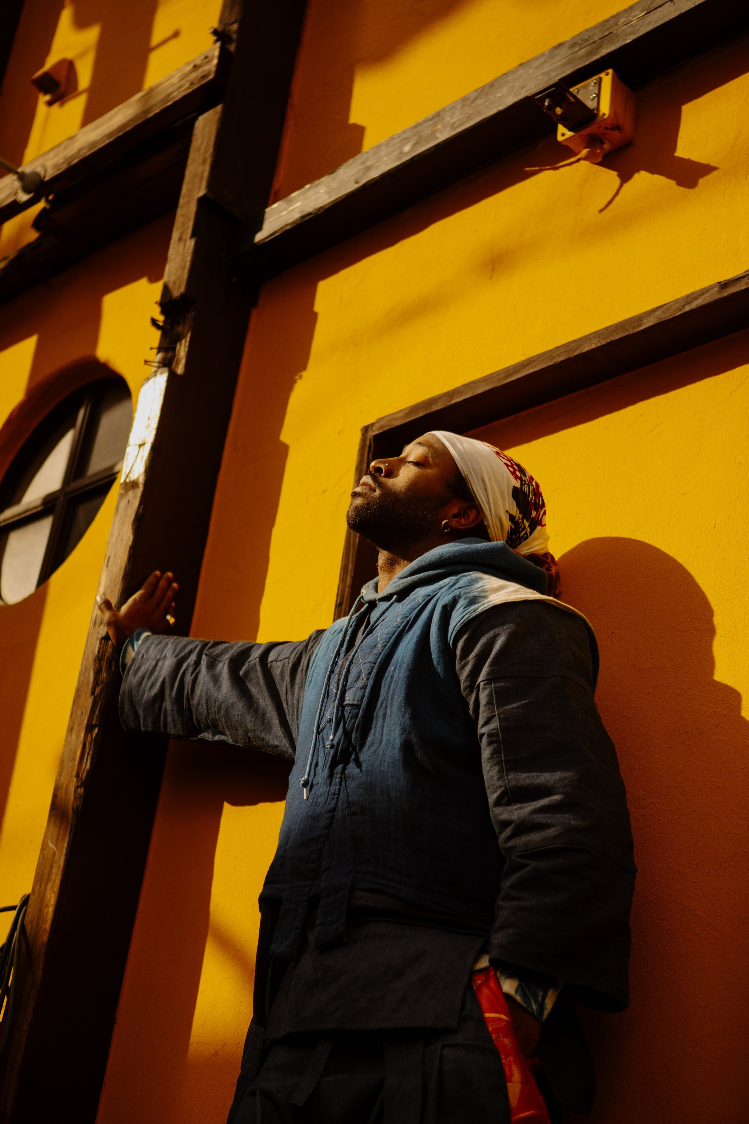 Solo editorial portrait of a man in a denim jacket and headscarf leaning against a sunlit yellow wall, showcasing solo portrait photography services