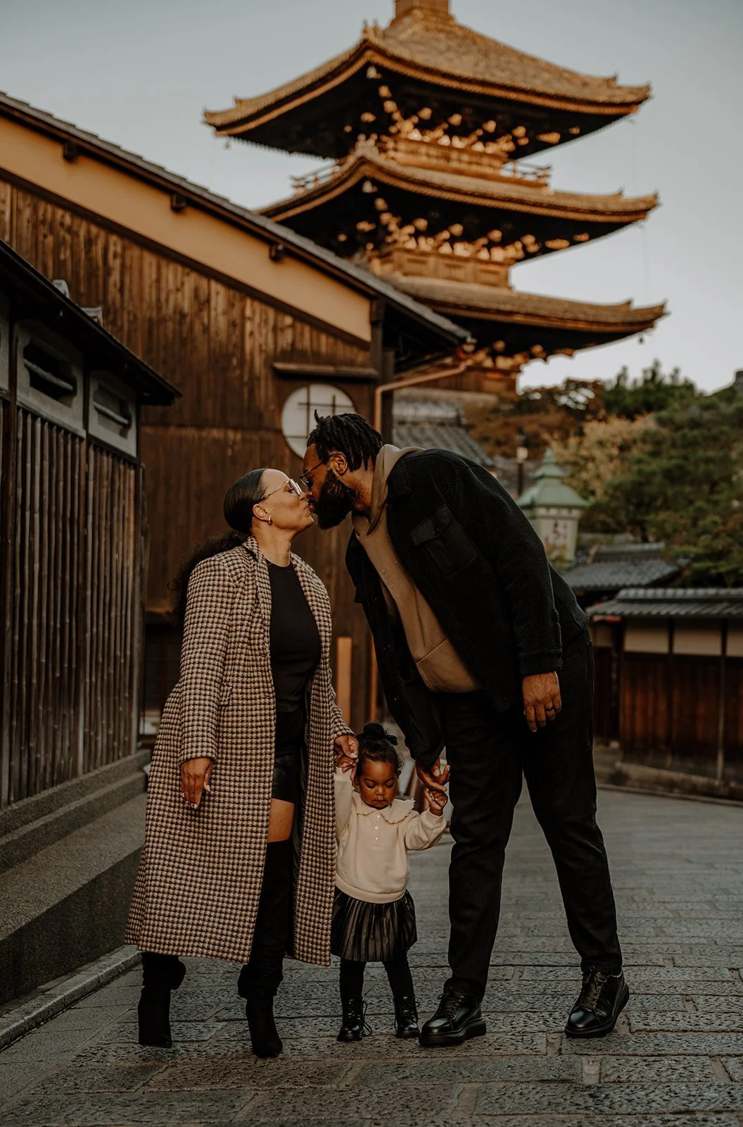 Family of four pausing on a Kyoto street lined with wooden shopfronts, a five-story pagoda rising behind them in daylight. 