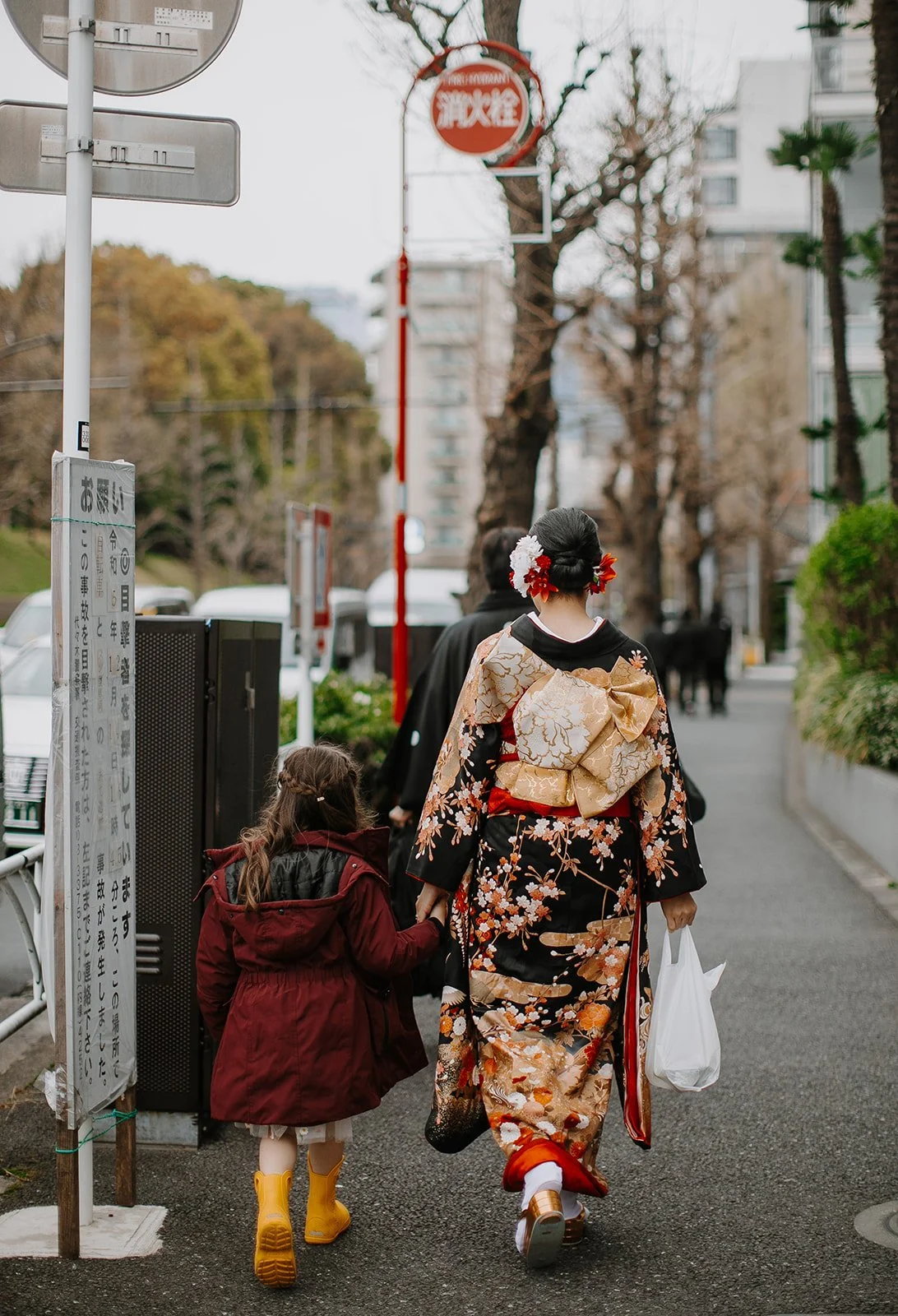 Woman in a formal kimono walks hand in hand with a child down a city street.