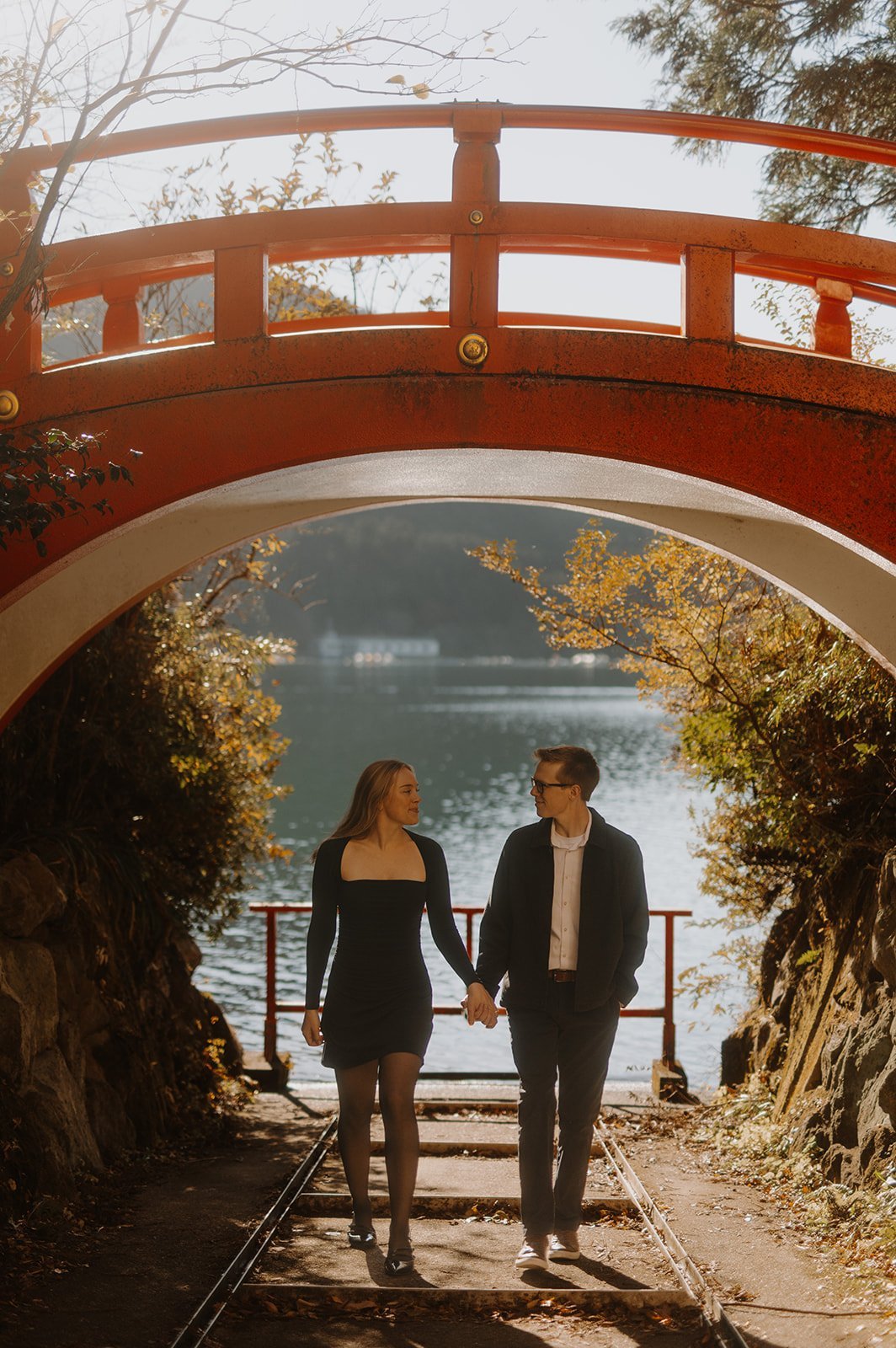 Couple walking hand in hand beneath a red arched bridge with Lake Ashi and autumn leaves behind them, Hakone