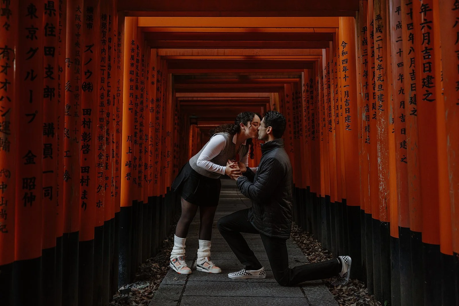 Man kneeling to propose inside the thousand vermillion torii gates at Fushimi Inari Shrine in Kyoto, the couple kissing as she says yes.