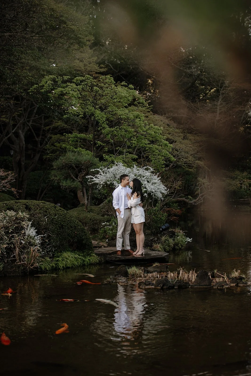 Couple in white outfits embracing beside a koi pond and blooming white blossoms in a Tokyo garden