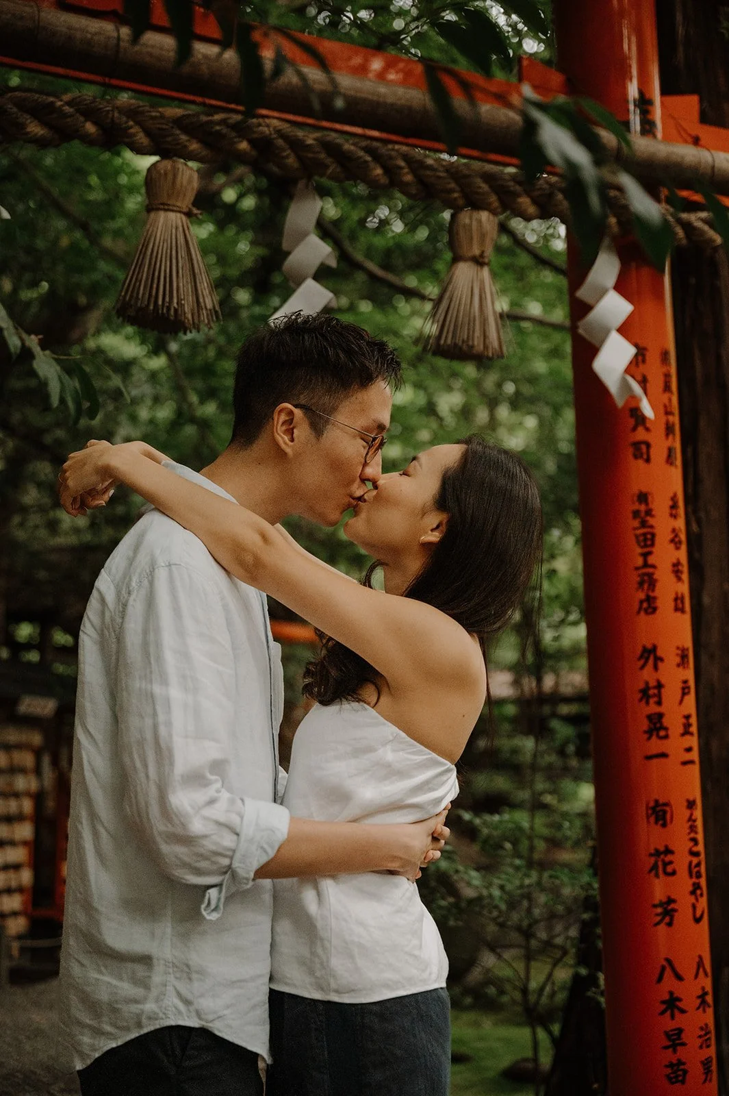 Couple kissing beneath a sacred shimenawa rope at the entrance to a red torii gate in a Kyoto shrine.