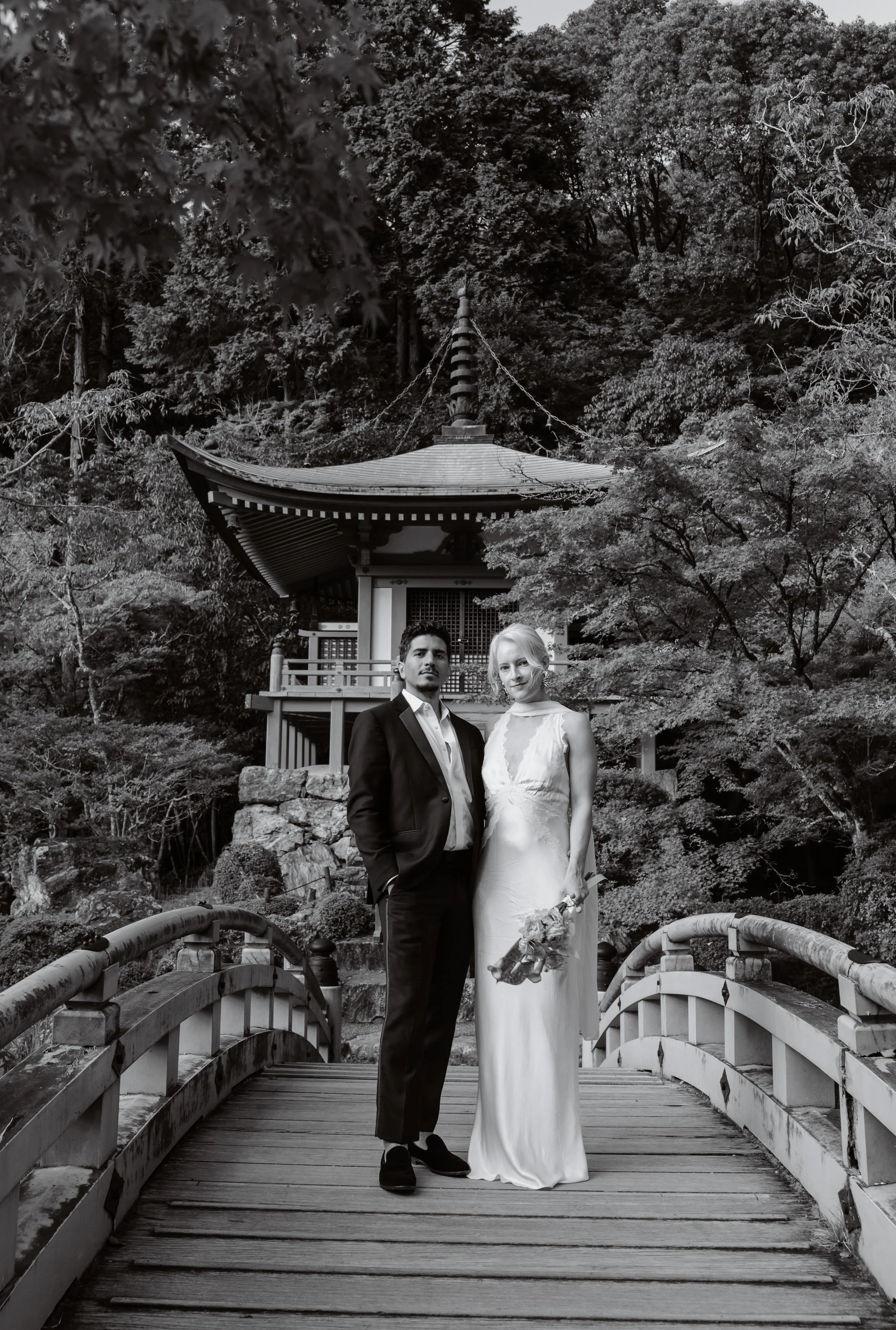 Black and white Kyoto pre-wedding portrait of a couple on an arched wooden bridge before a two-tiered Japanese pagoda and forested garden.