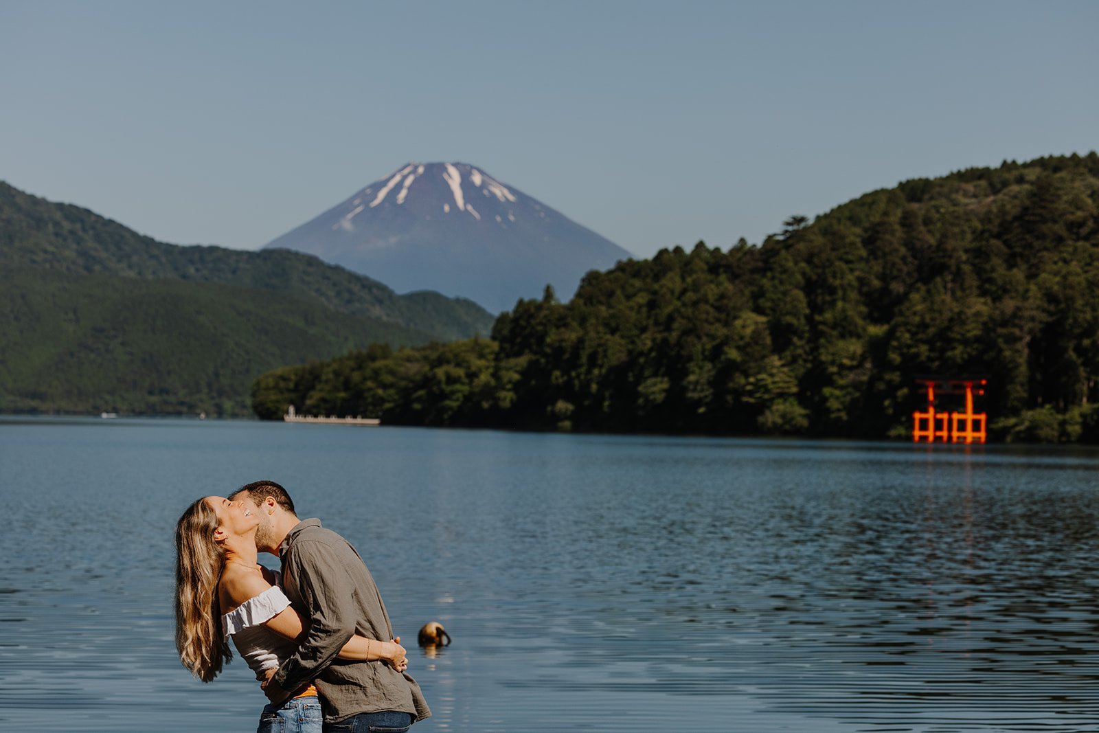 Engaged couple kissing on the shore of Lake Ashi with Mount Fuji and the floating torii gate visible in the distance, Hakone, Japan