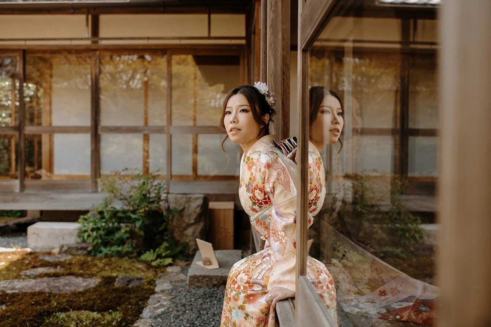 Woman in a floral kimono leaning against a glass door beside a traditional Japanese garden, reflection visible in the glass