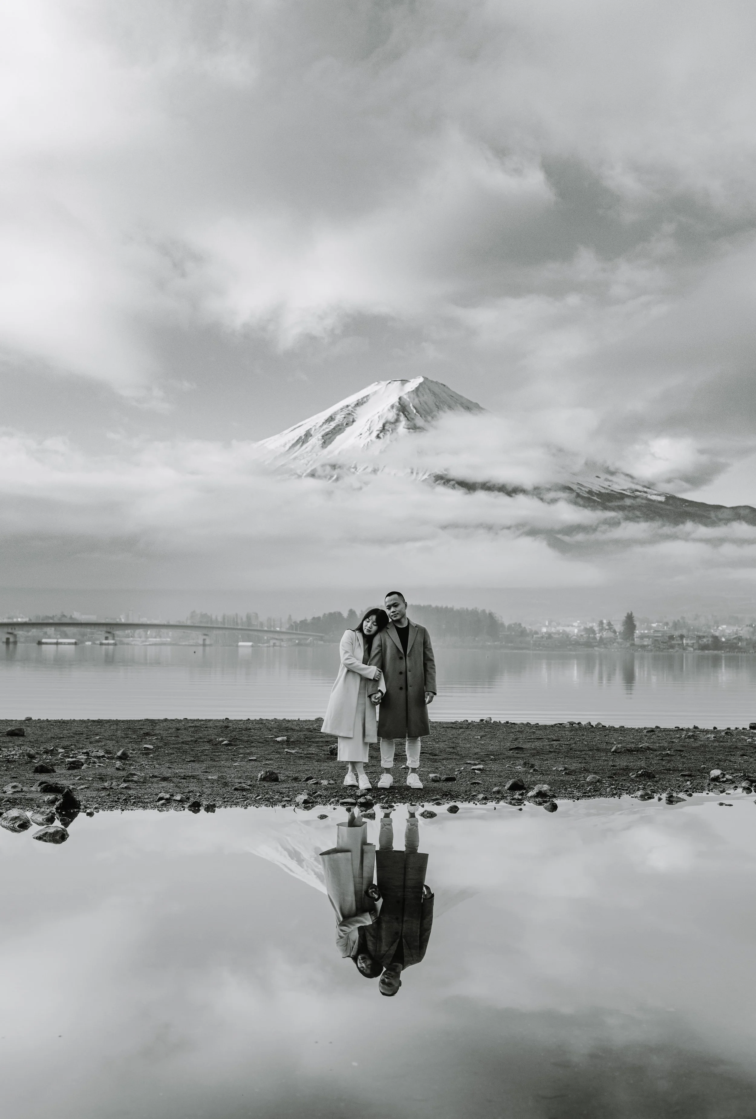 Black and white portrait of a couple standing lakeside with their reflection and Mount Fuji in the background