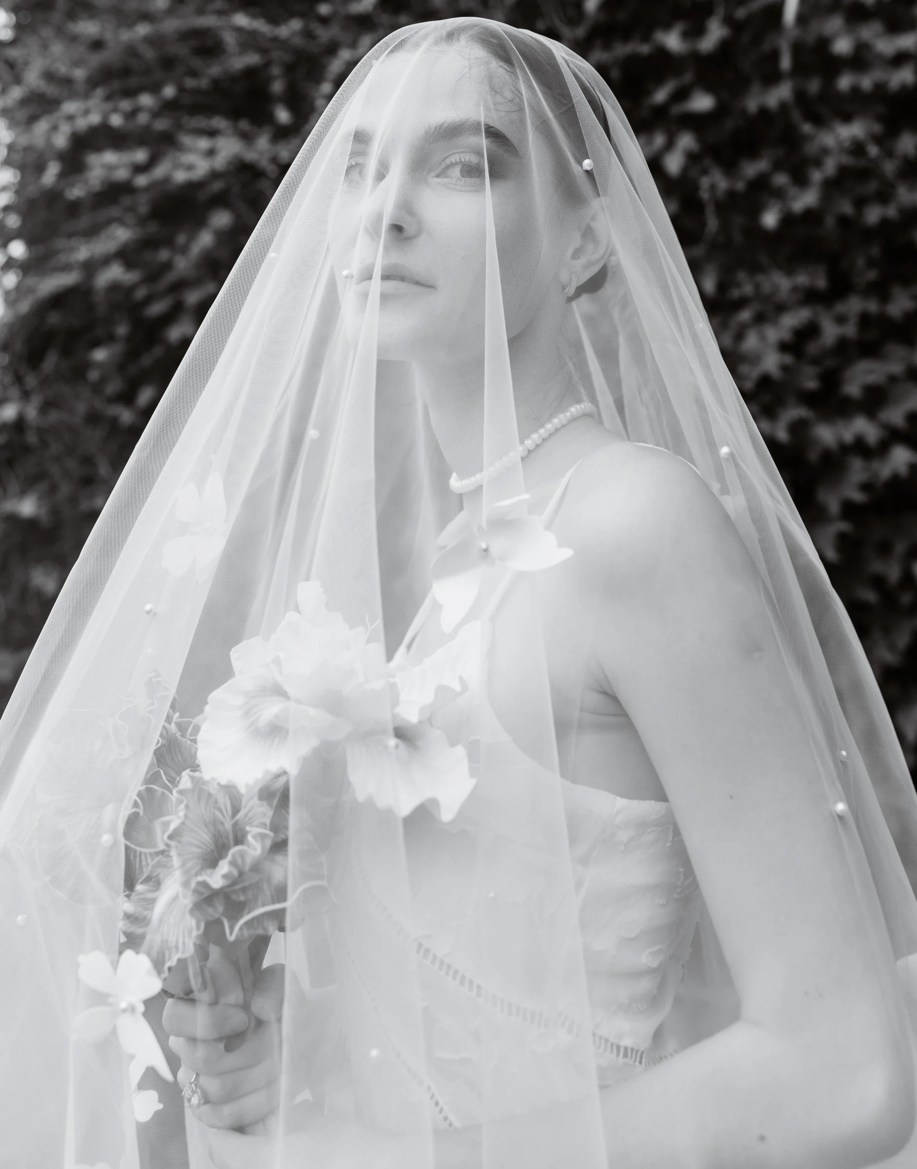 Black and white portrait of a bride beneath a sheer pearl-detailed veil holding a floral bouquet