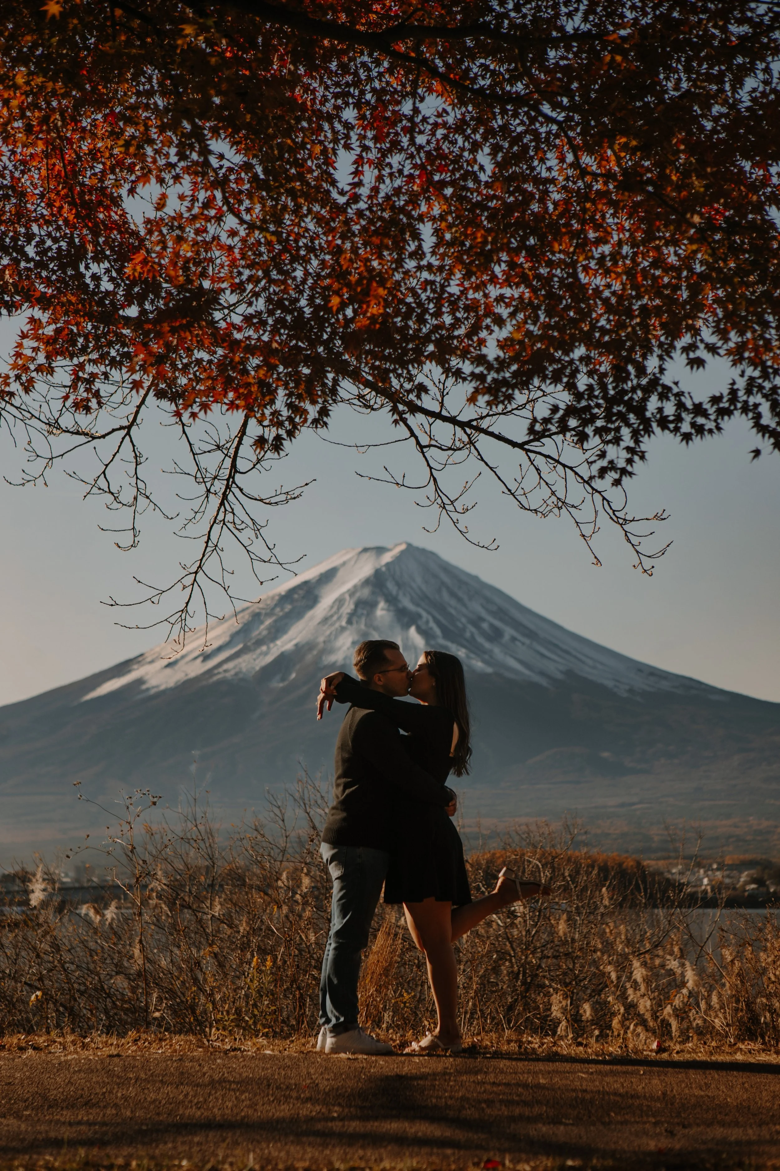 Couple sharing a kiss under a red autumn maple tree with Mount Fuji visible in the distance