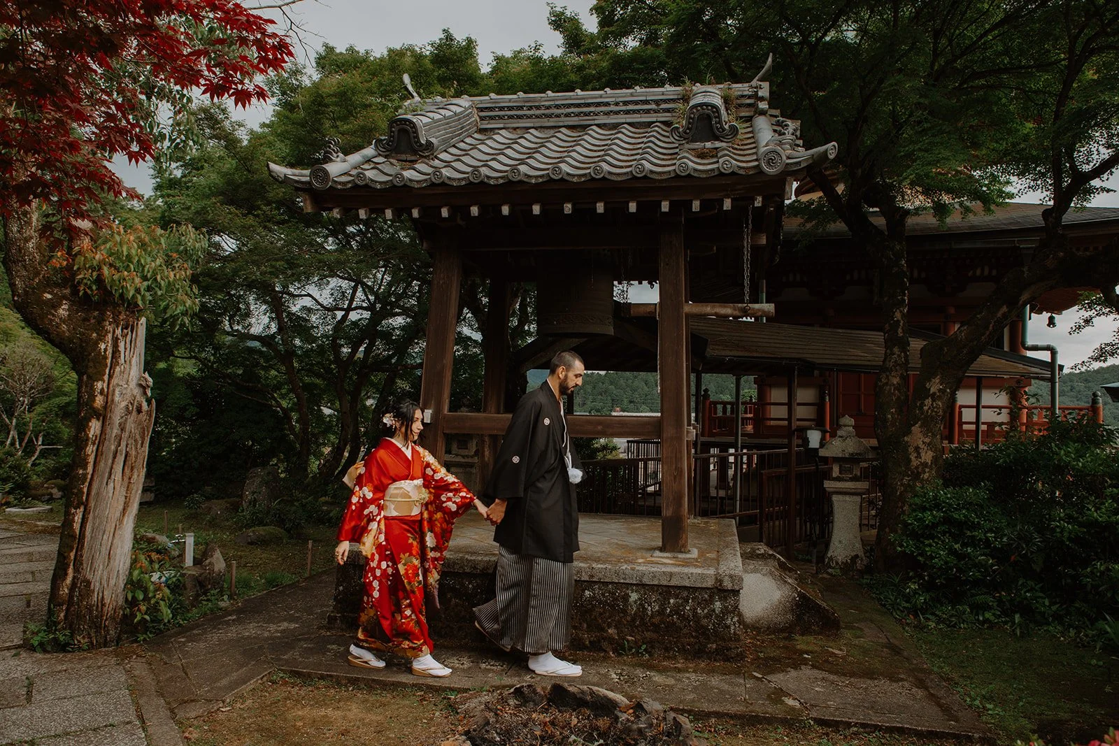 Couple in kimono walk hand in hand past a wooden temple bell tower overlooking the Kyoto hills.