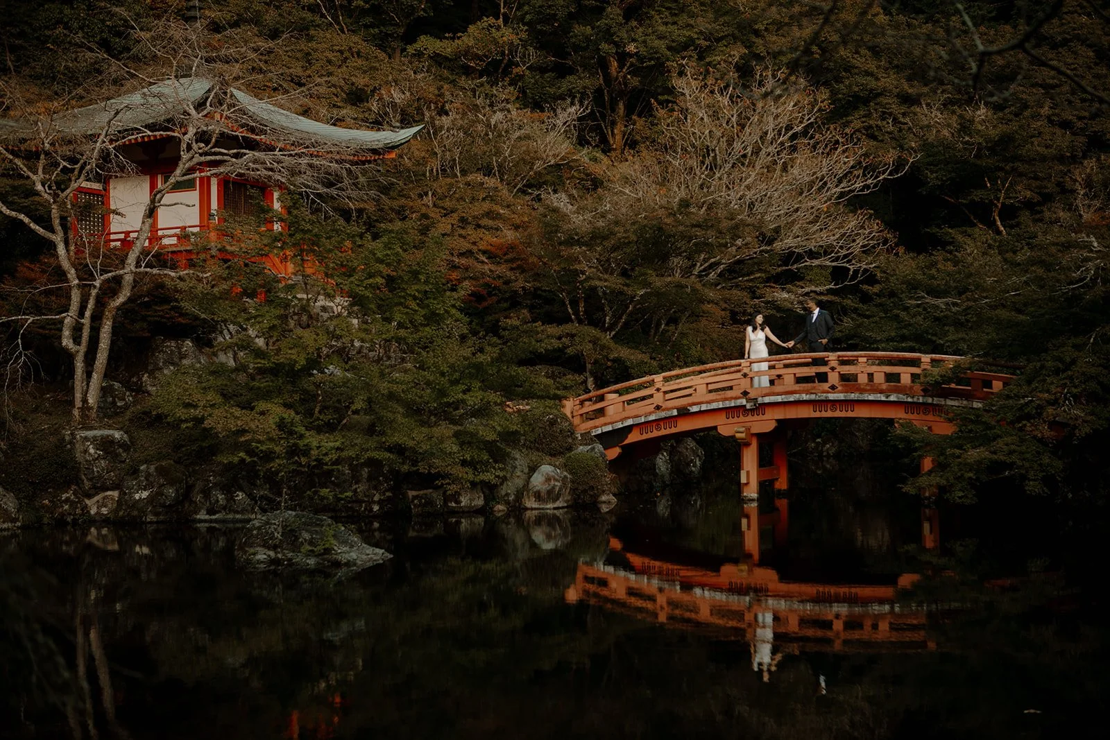 Bride and groom stand on the red temple bridge at Daigo-ji, mirrored in the still water below.