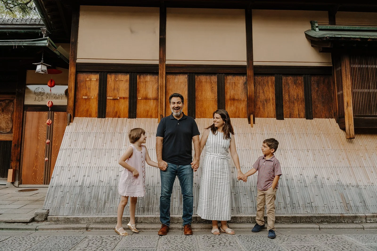 Family of four holding hands and laughing together in front of a traditional wooden ryokan facade, showcasing family portrait photography services