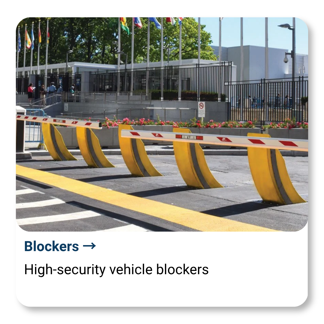 High-security vehicle blockers in front of a gated building with flags, a flower bed, and people outside on a sunny day.