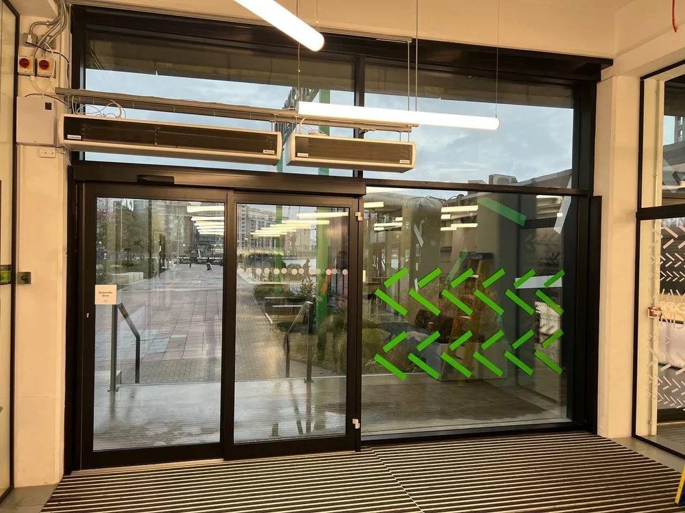 Glass storefront door with green arrow decorations on the inside window, showing an outdoor walkway and some buildings reflected in the glass.