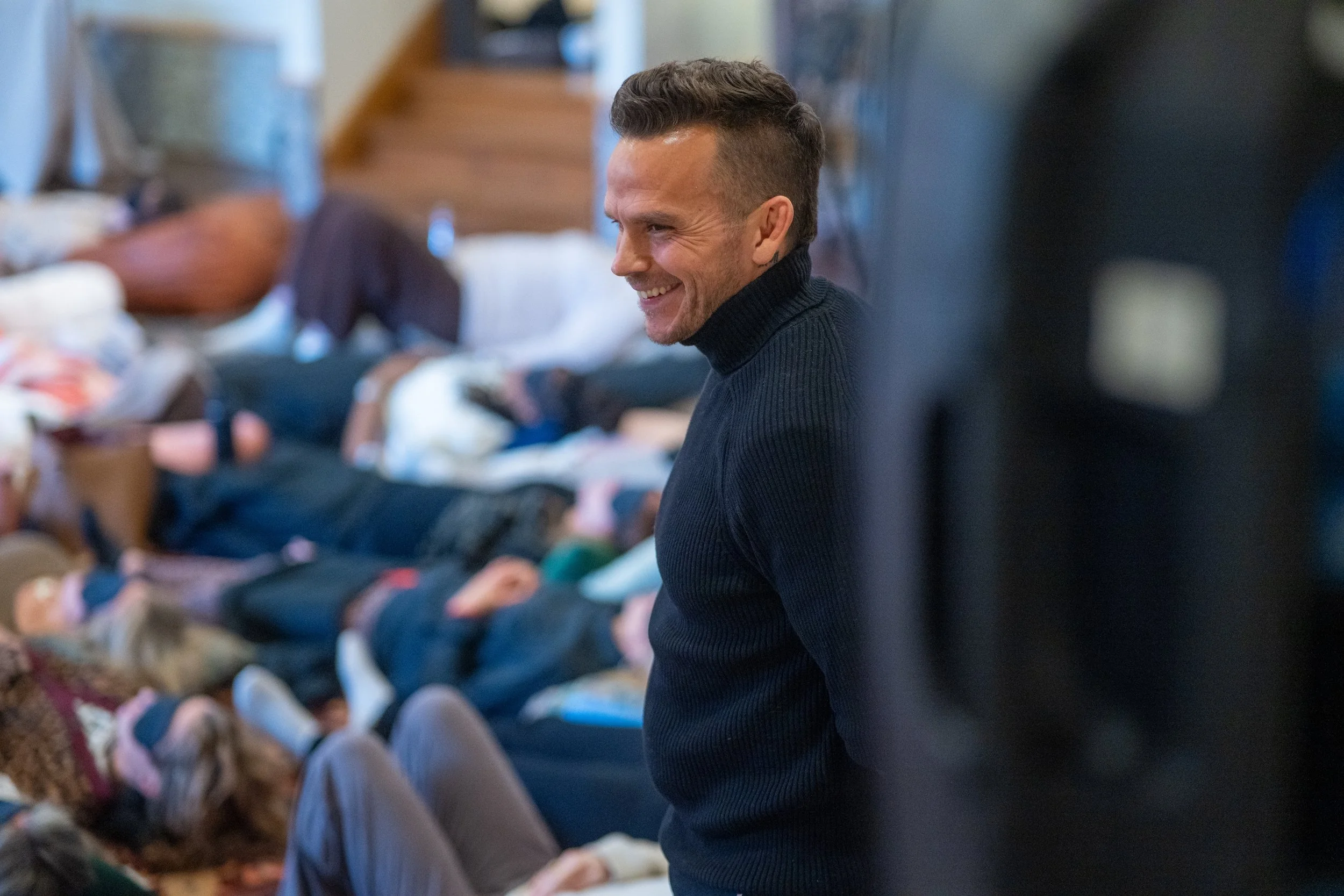 A smiling man in a dark turtleneck stands in a crowded room with many people lying on the floor.