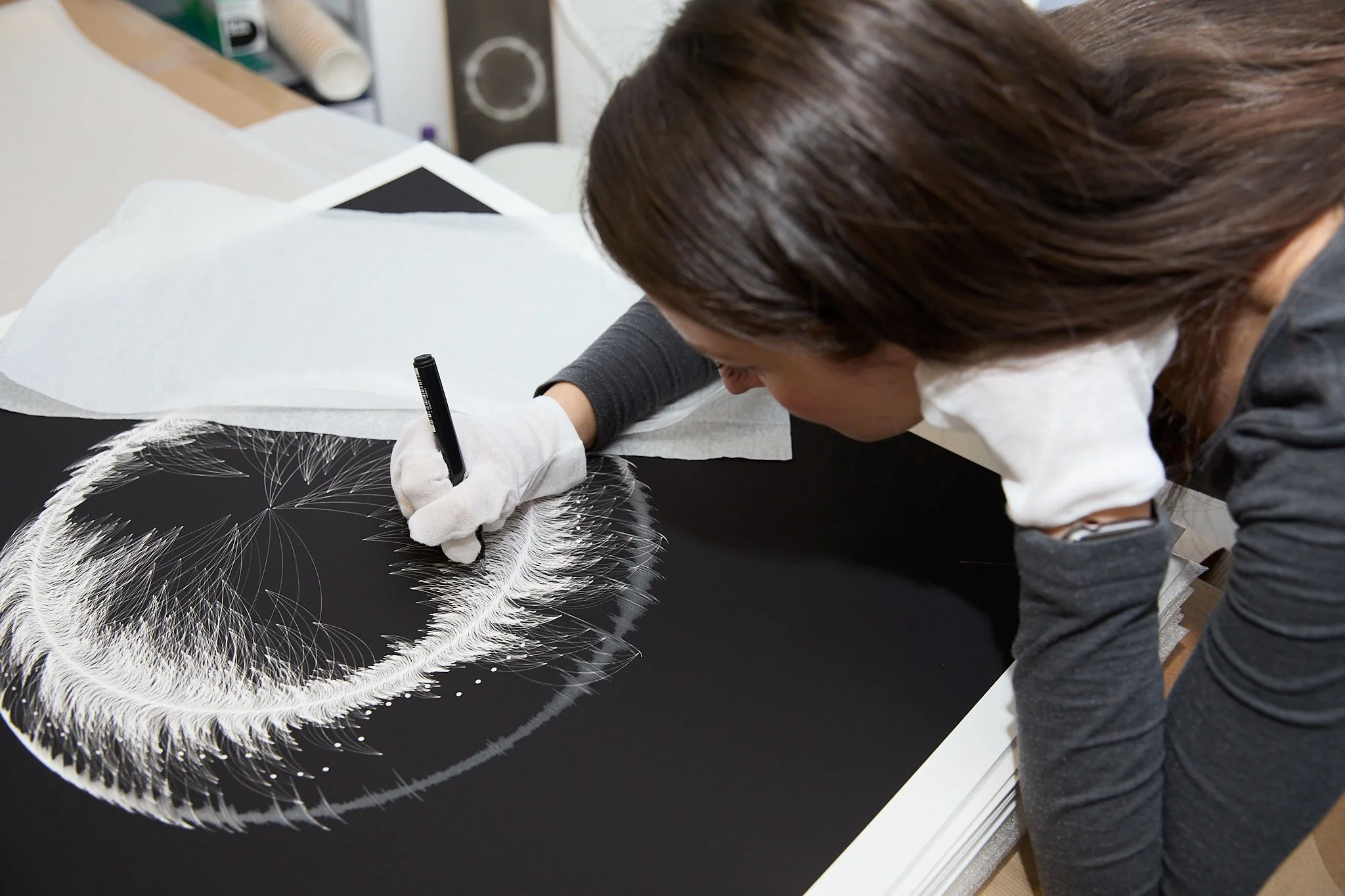Person creating a detailed white feather illustration on black paper using a marker, surrounded by protective paper sheets.