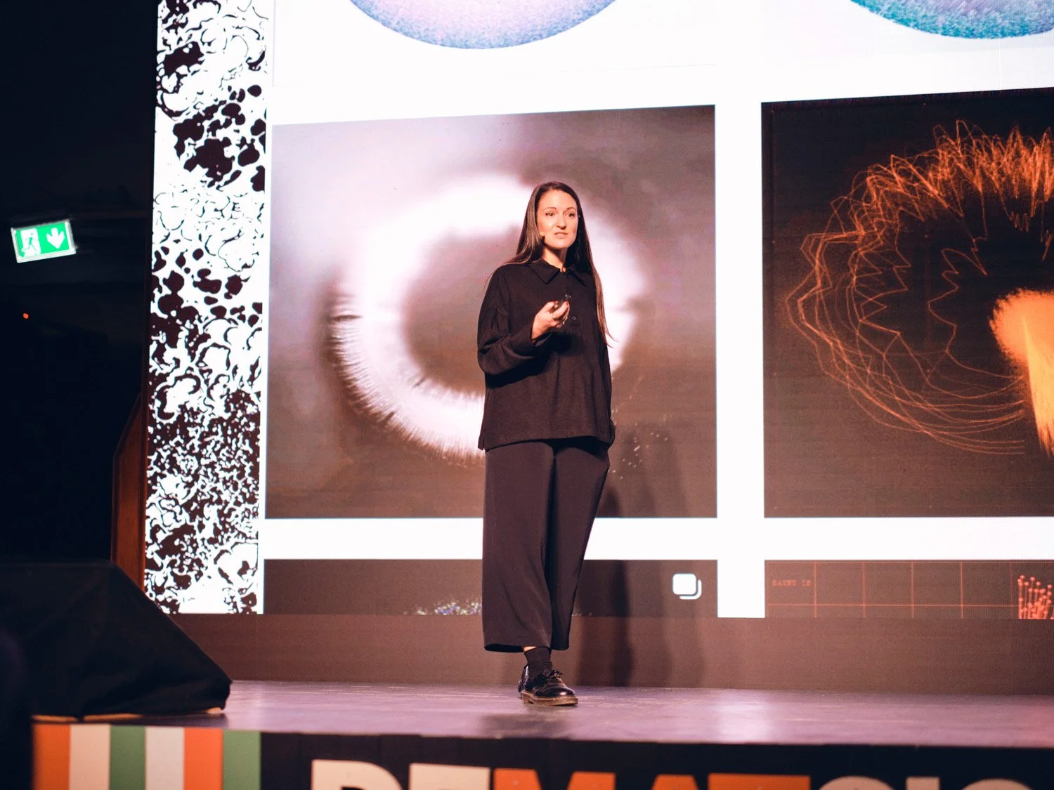 A woman in black clothing giving a presentation on a stage with digital screen backgrounds.