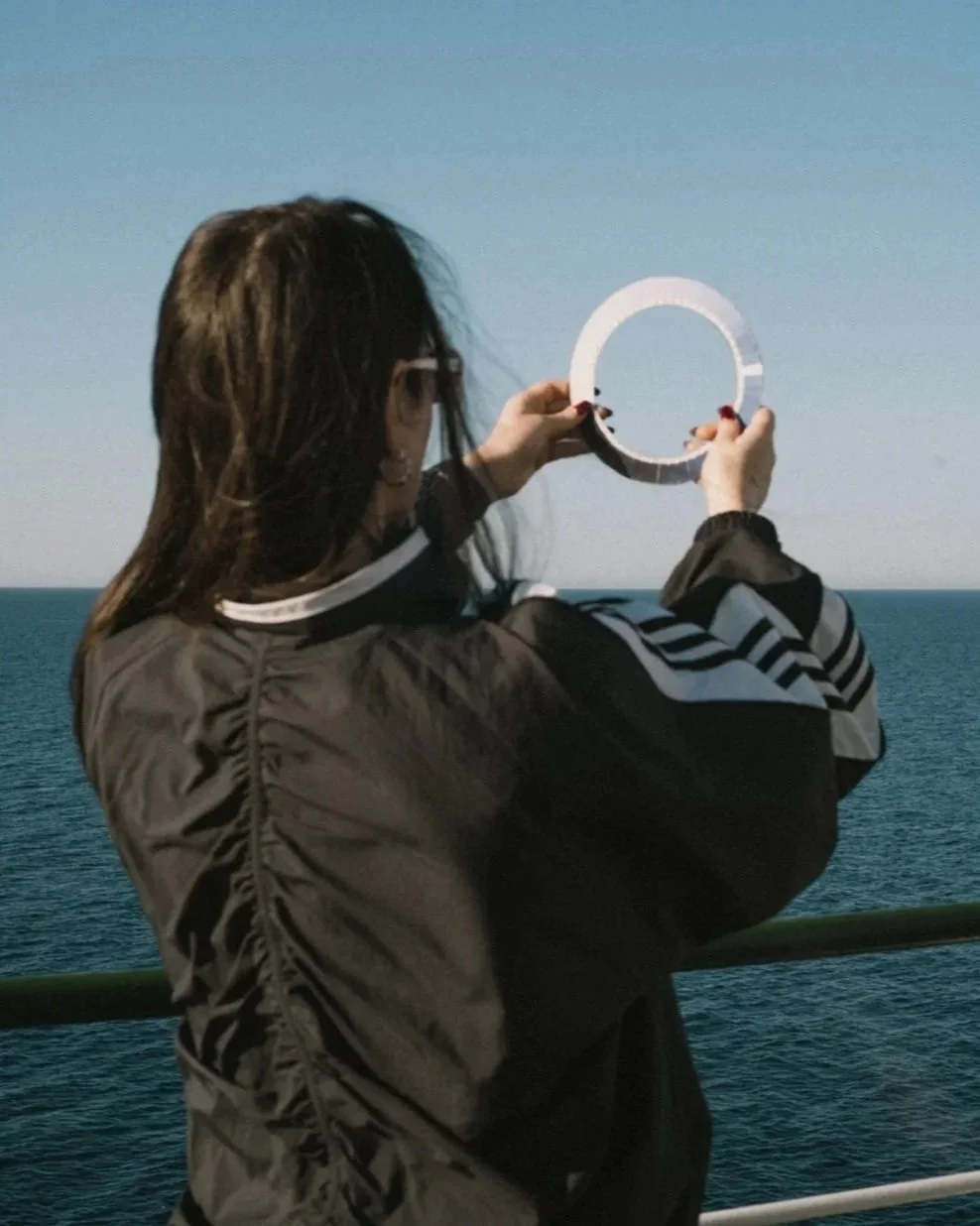A woman with dark hair and glasses, wearing a black and white jacket, is holding up a round mirror on a boat with an ocean background.
