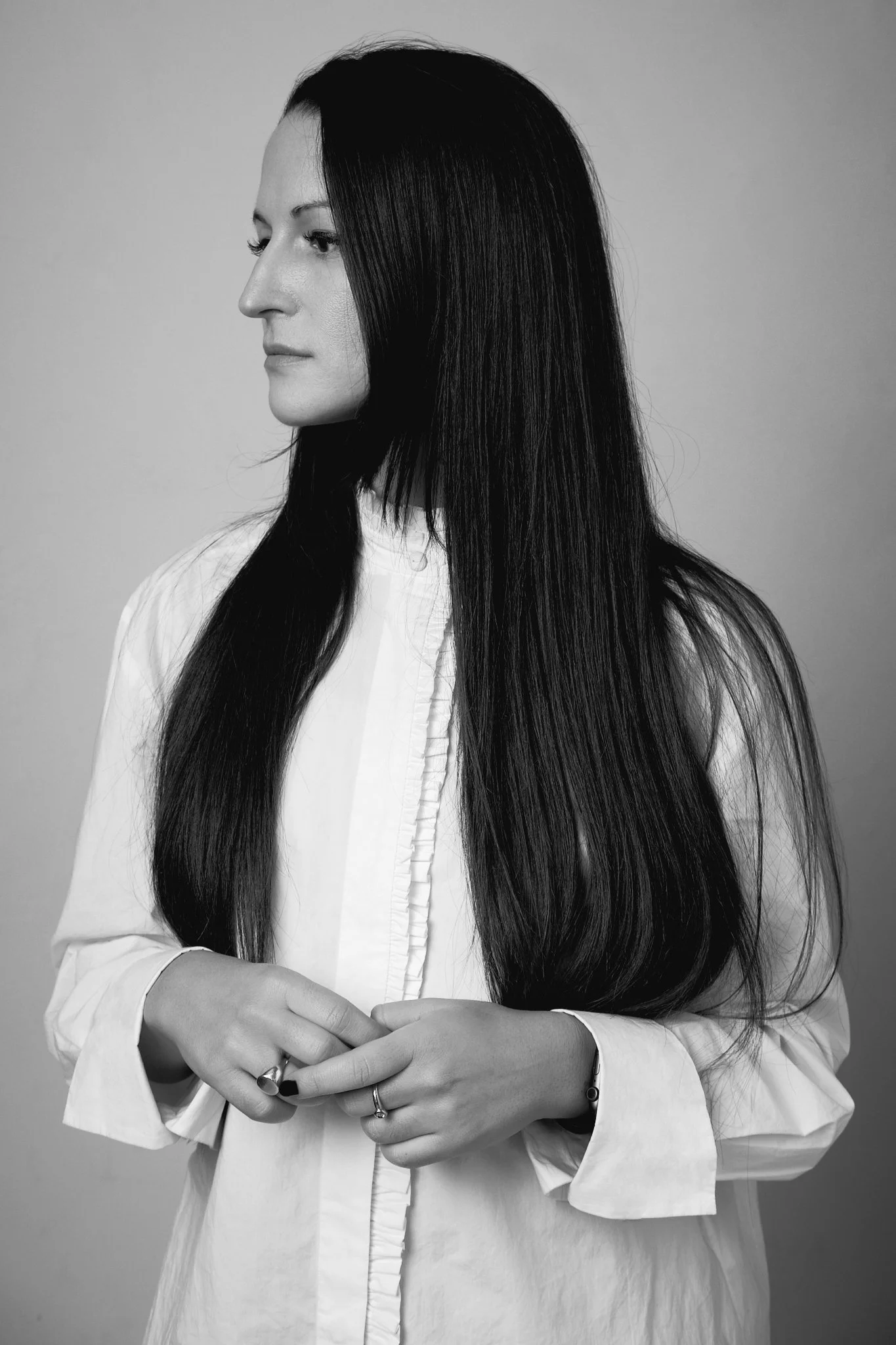 Black and white photo of a woman with long dark hair, wearing a white shirt with gathered front details, standing against a plain background, with her hands gently clasped at waist level.