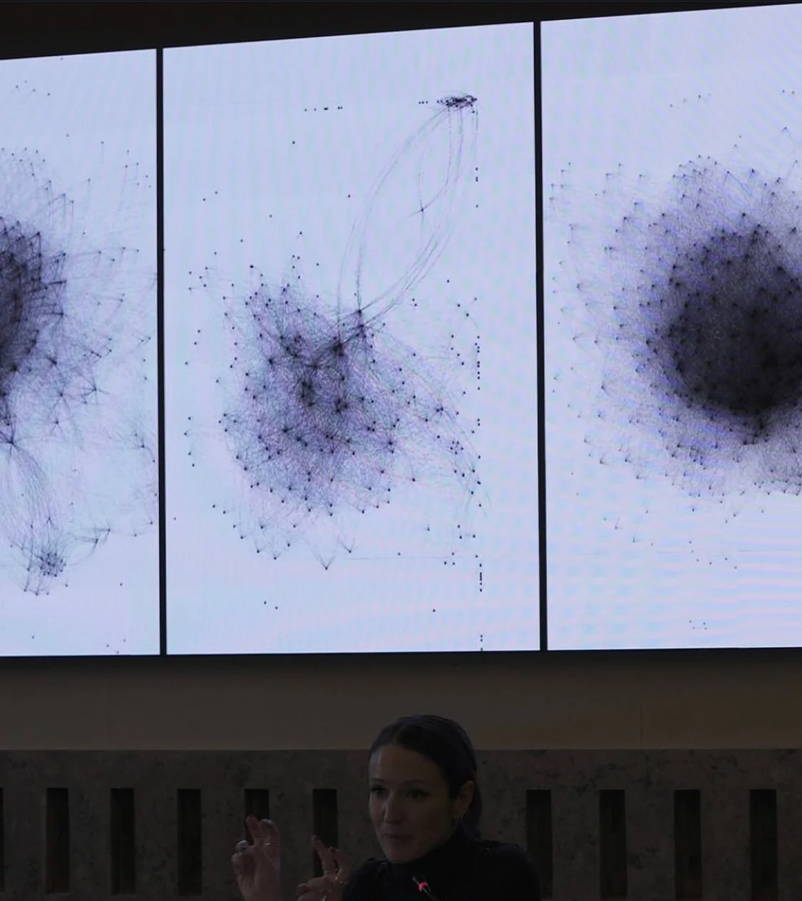 A woman standing in front of a large digital screen displaying complex interconnected node diagrams or network visualizations.