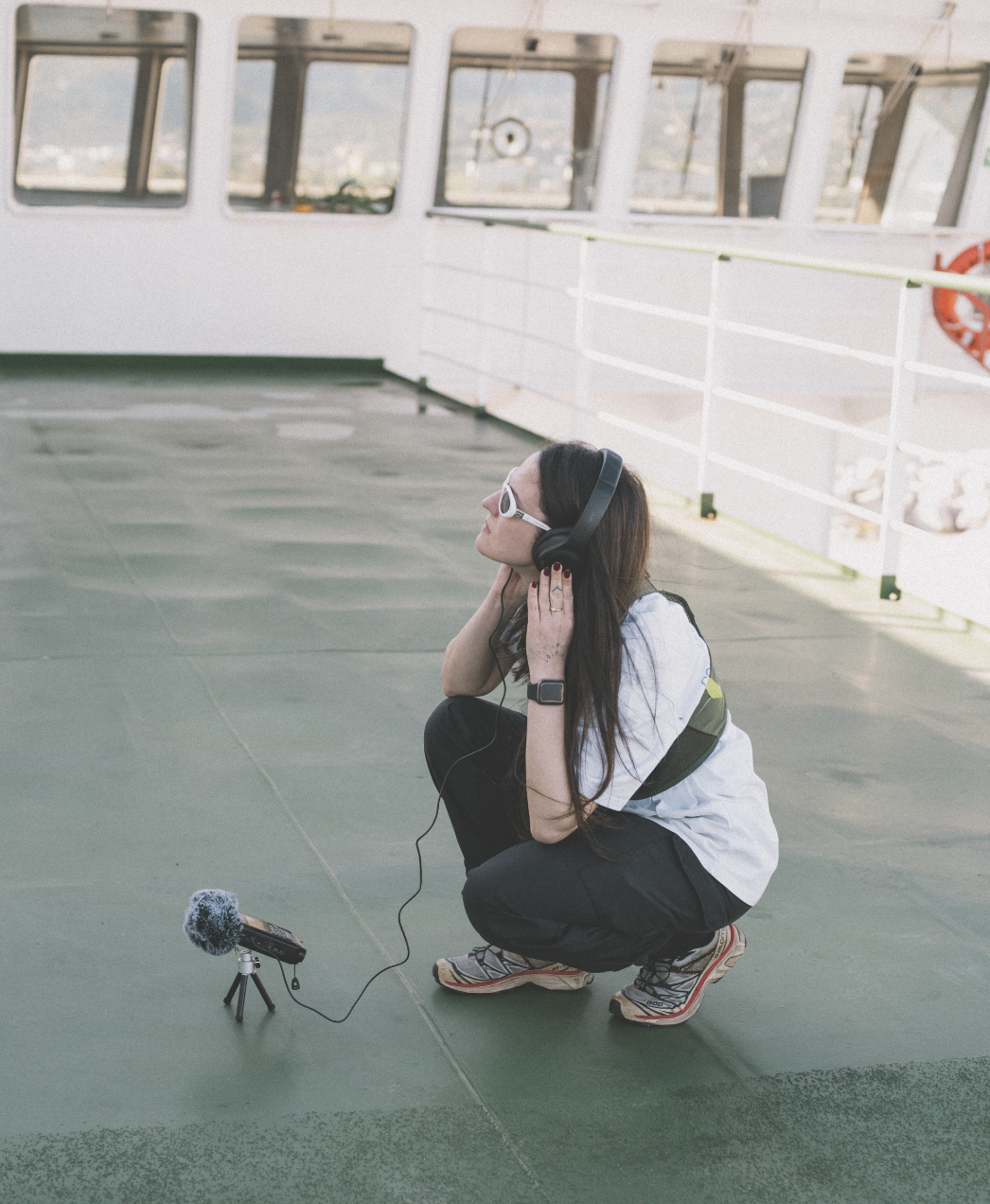 A woman with long dark hair wearing sunglasses, headphones, a white t-shirt, black pants, and sneakers crouches on a deck with a green surface, near the railing of a boat or ferry. She is wearing an Apple Watch and has a small recording device with a