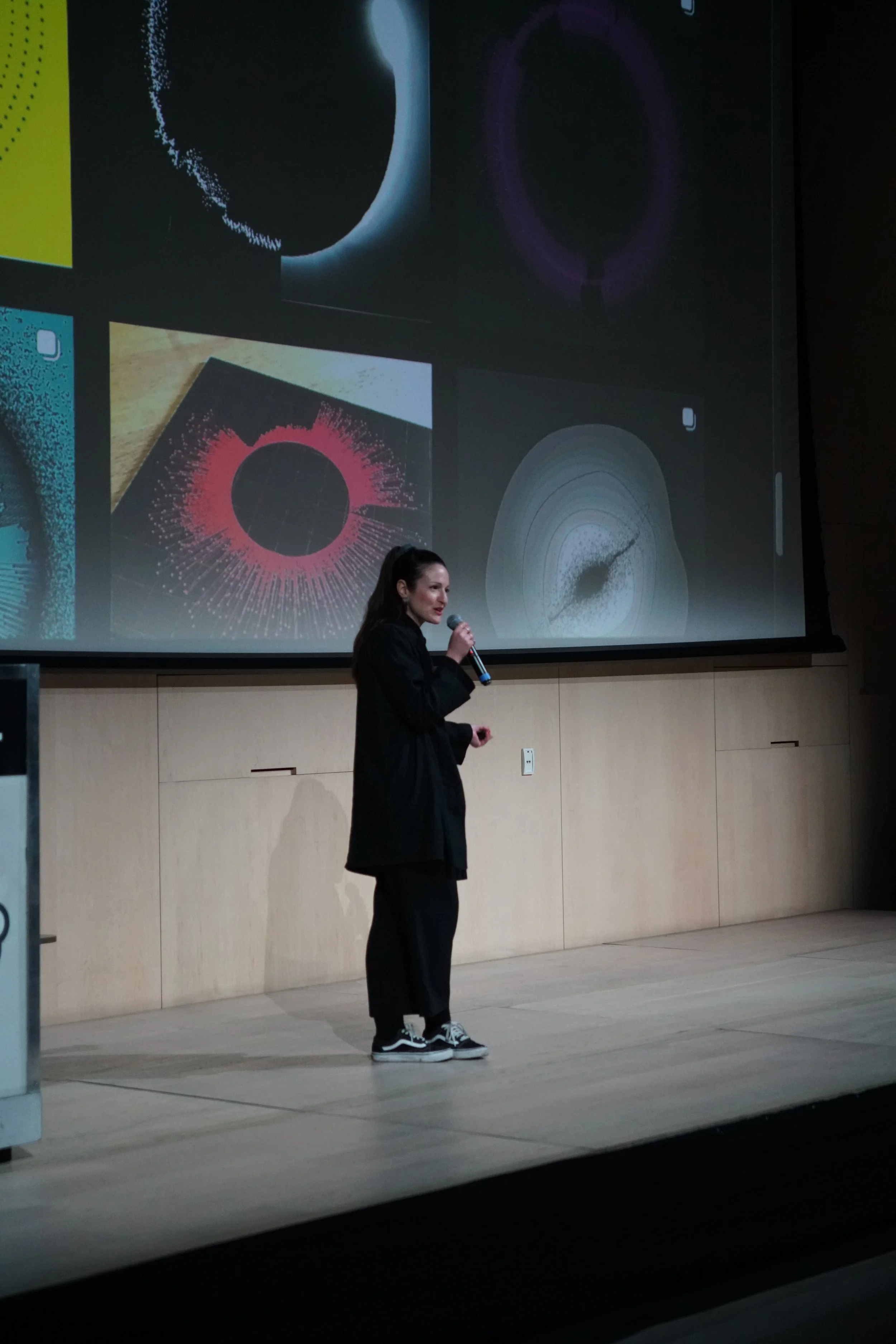 A woman with long dark hair, wearing a black outfit and sneakers, speaking into a microphone on a stage with a large screen behind her displaying various abstract colorful images.