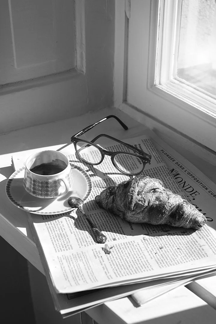 Stämningsfull svartvit fotokonst med croissant, kaffe, glasögon och tidning i ett fönster. Ett minimalistiskt fotografi som fångar en lugn och elegant morgonstund.