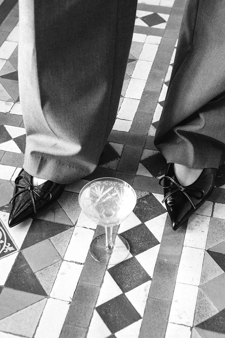 Svartvit fotokonst av eleganta skor och vida byxor på ett dekorativt mosaikgolv med ett glas i förgrunden.