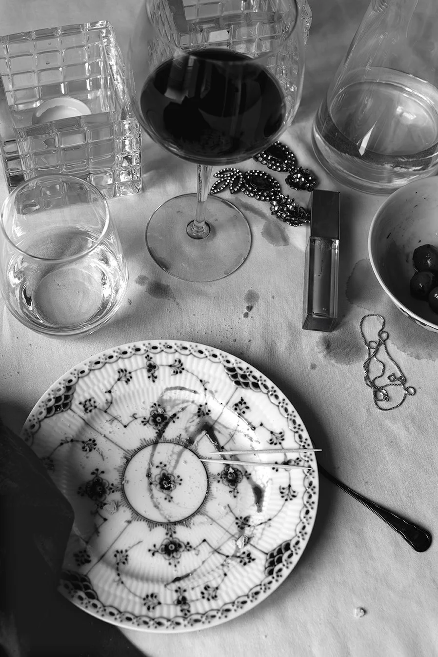 Svartvit fotokonst i stillebenstil med vinglas, karaff, glas, oliver, smycken och dekorativ tallrik på ett elegant dukat bord.