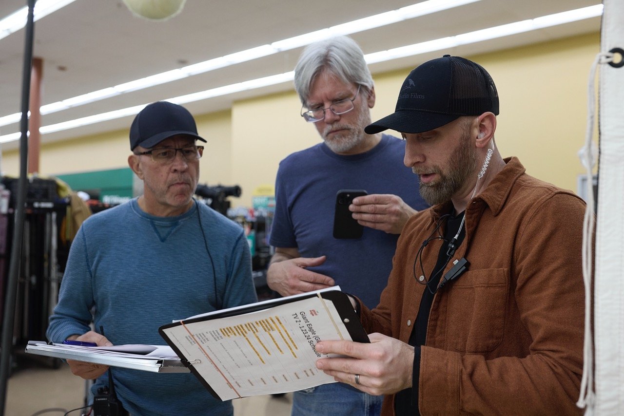 Director, DP and AD reviewing shot list on set, in a brightly lit room with filming equipment in the background.
