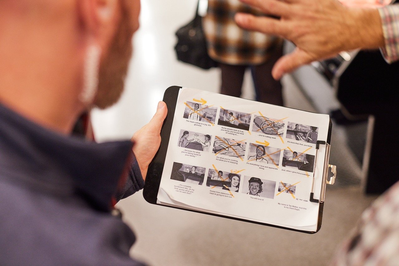 Director holding a clipboard with a storyboard containing illustrations and text. Another person's hand is pointing at the storyboard. The setting appears to be indoors, in a busy environment with people present.