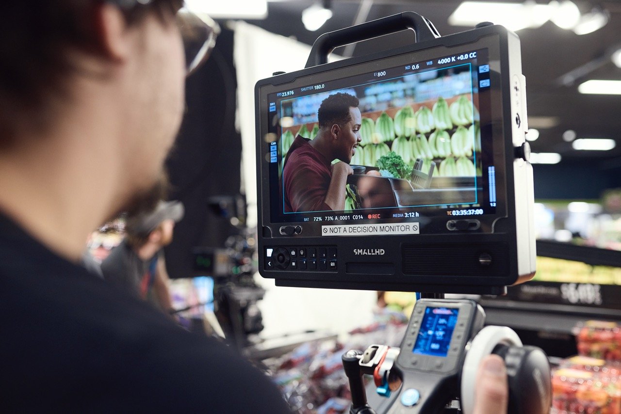 Camera crew filming a man in a supermarket, monitor shows man with shopping cart in fruit aisle.