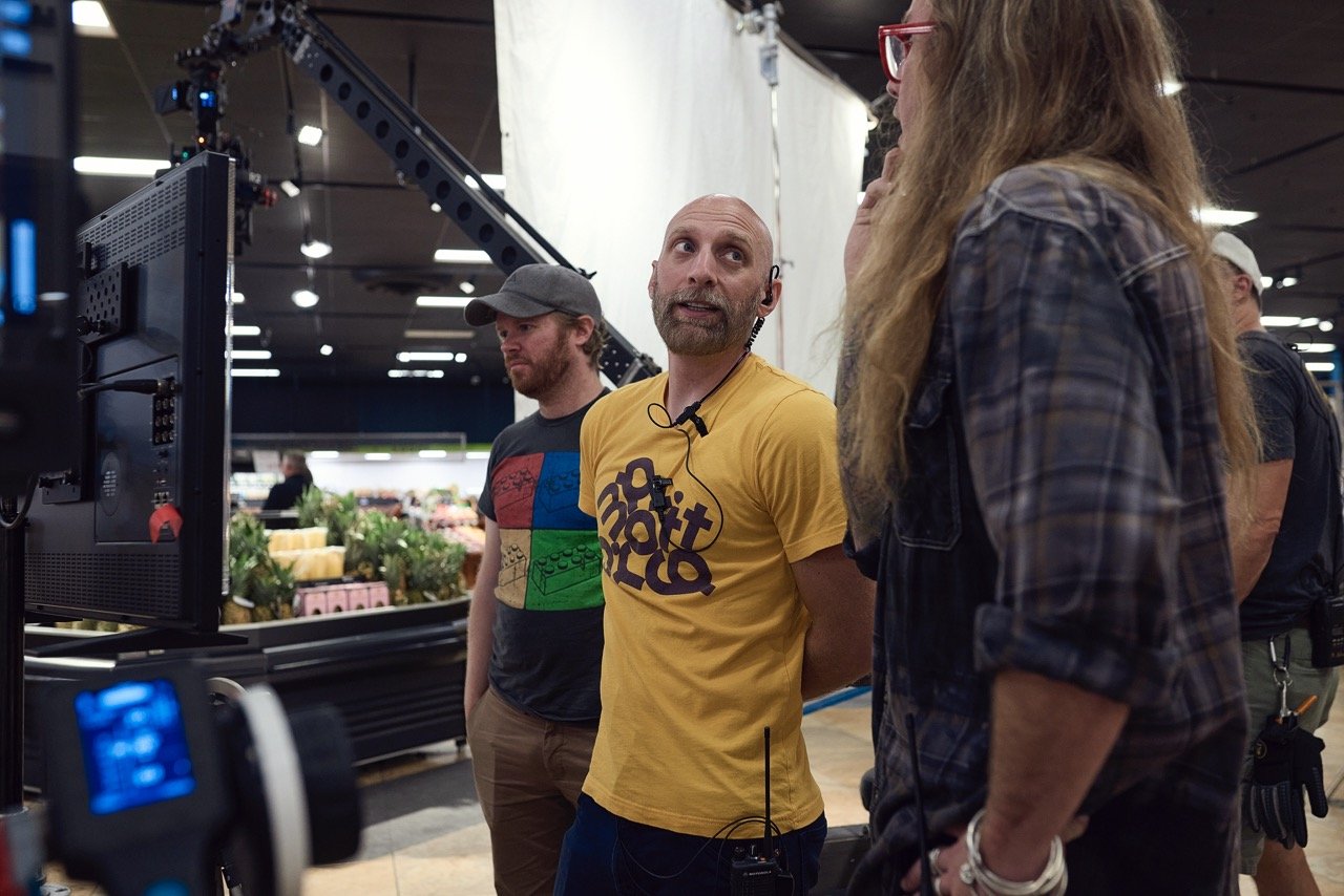 Production crew members on a set with lights and filming equipment. They are discussing something near a monitor.