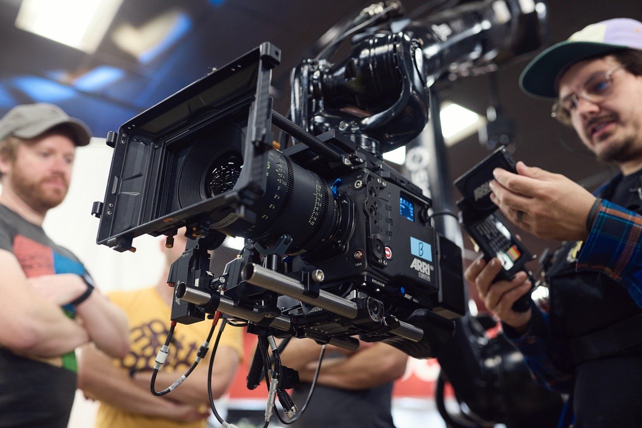 Close-up of a professional film camera setup with Arri branding, surrounded by individuals adjusting equipment in a studio setting.