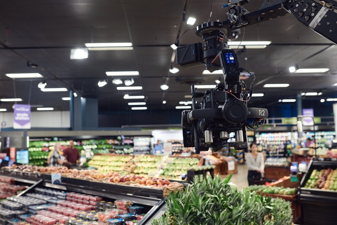 Film camera setup on crane arm in grocery store produce section with fruits and vegetables.