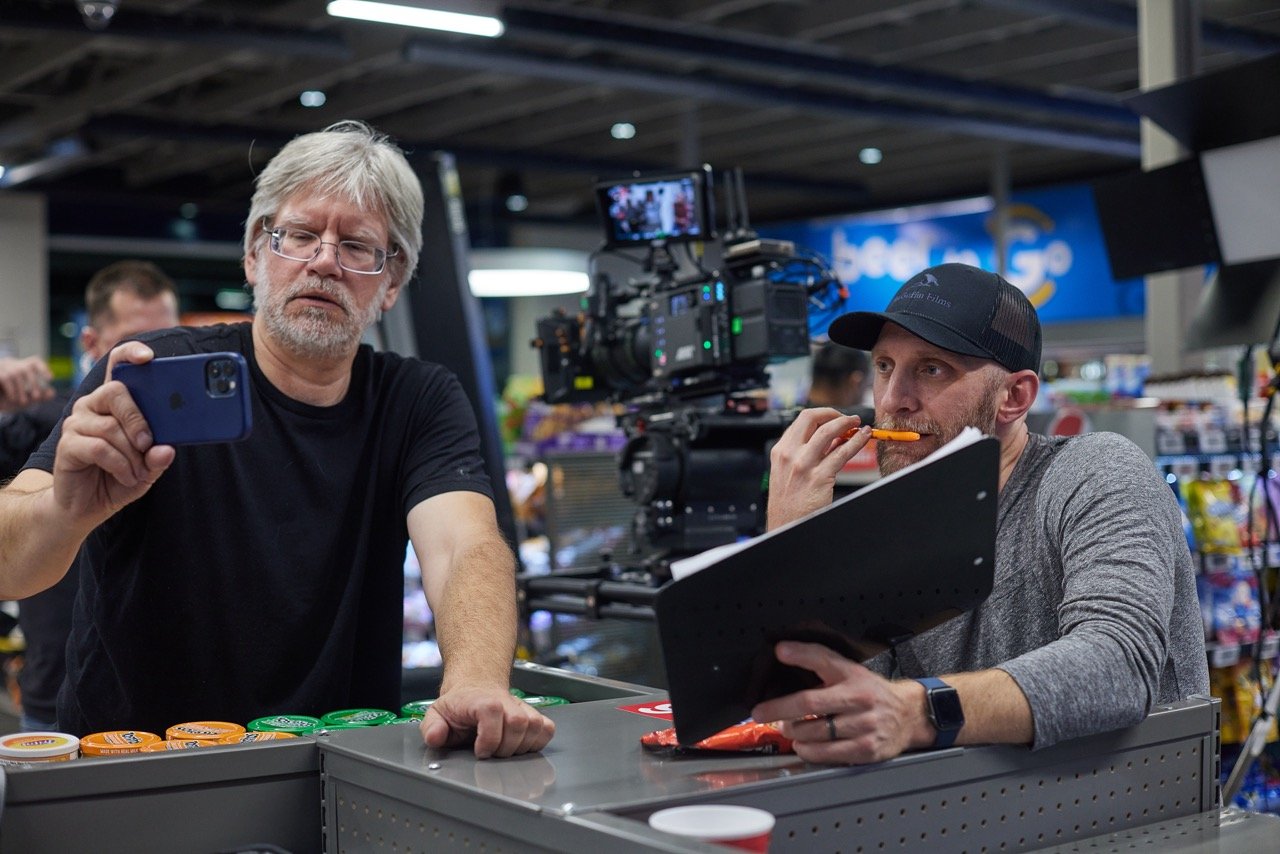 Director and director of photography on a commercial film set, one holding a smartphone, the other appearing to review notes with a clapperboard nearby. A camera is mounted on a rig beside them.