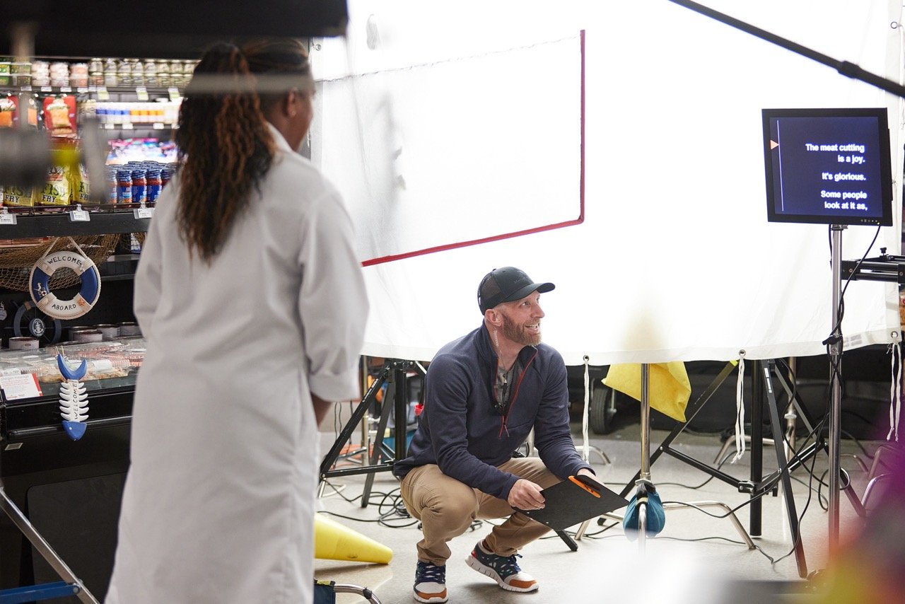 Director crouching with a clipboard in a filming set inside a grocery store, with a woman in a white coat in the foreground and a teleprompter in the background.