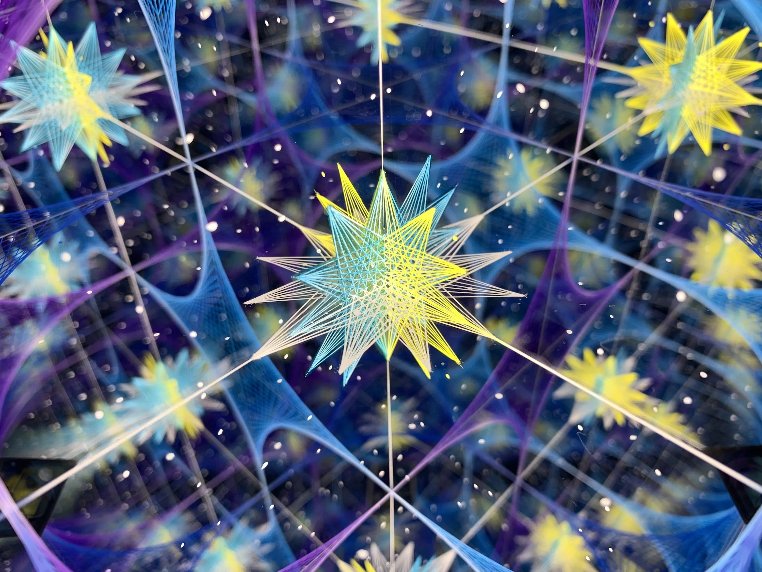 A digital art image featuring multiple colorful, star-shaped, spiky geometric structures interconnected with thin lines. The stars are predominantly yellow and blue with accents of purple and white, set against a dark cosmic background with small white dots resembling stars.