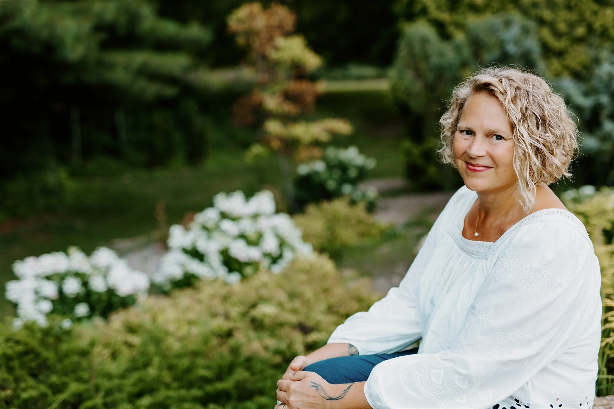 A woman with curly blonde hair, wearing a white blouse, sitting outdoors in a garden with greenery and white flowers, smiling at the camera.