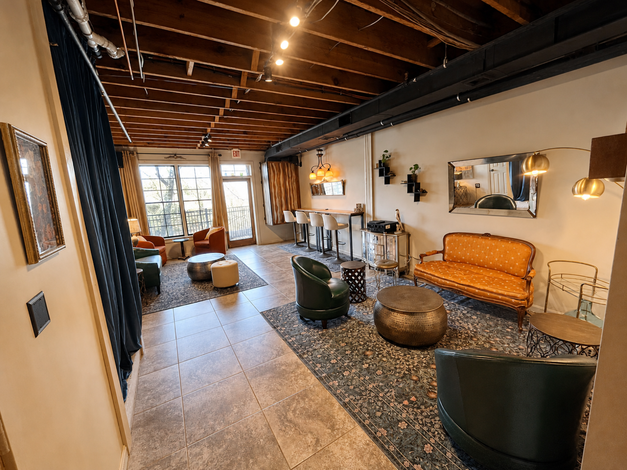 A cozy indoor lounge area with large windows, wooden ceiling beams, various chairs, a sofa, a mirror, and ambient lighting.