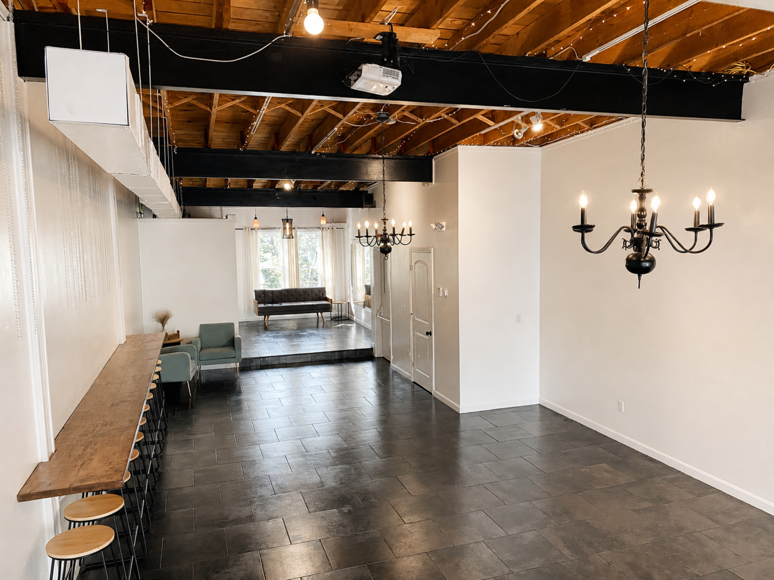 Interior of a room with a wooden ceiling, ceiling lights, a chandelier, large windows with white curtains, white walls, dark floor tiles, a seating area with a brown tufted bench, green armchairs, and a long wooden bar table with stools along the wall.
