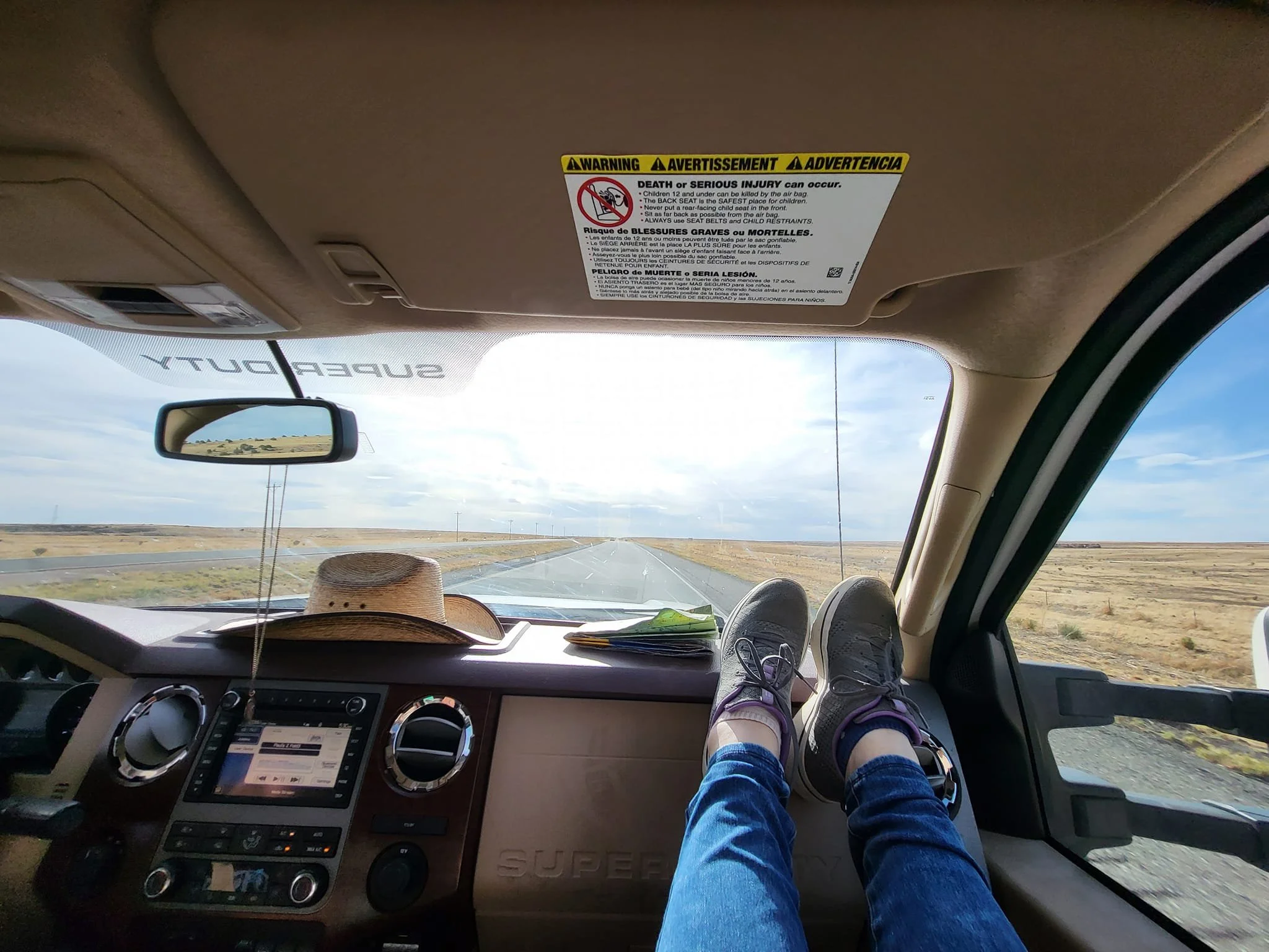 Inside a vehicle, a person wearing blue jeans and purple sneakers is resting their feet on the dashboard, facing a road visible through the windshield. A straw hat and some papers are on the dashboard. The rearview mirror shows a landscape of flat land and power lines. A warning label with safety instructions is on the sun visor.