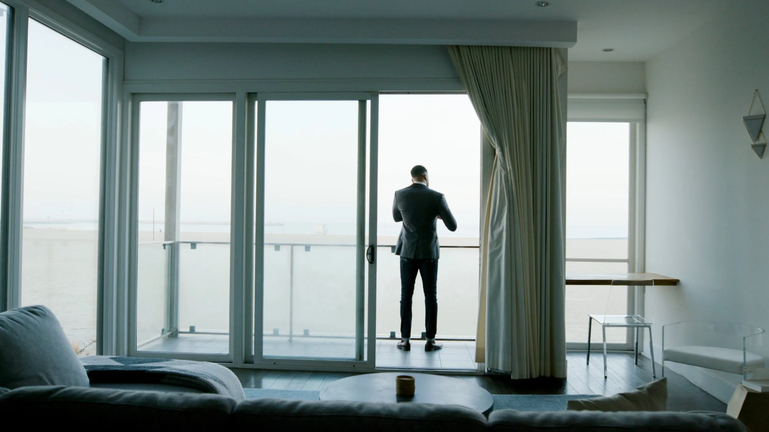 A man in a suit standing on a balcony, looking out over the water, inside a modern living room with large glass doors and minimal decor.