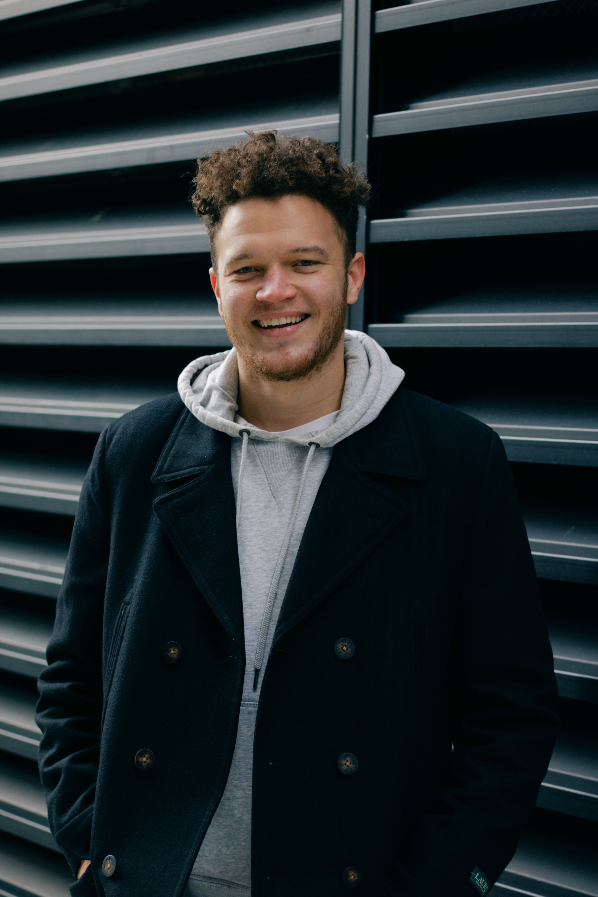 Young man smiling, standing outdoors against a modern metal wall with horizontal louvers, wearing a dark coat and a gray hoodie.