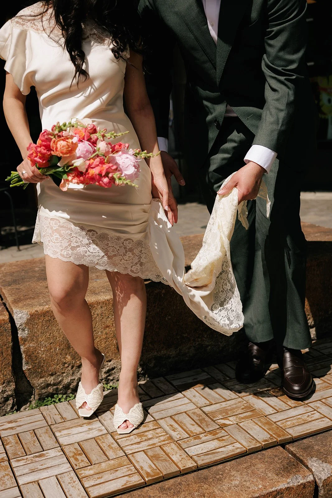 Partially visible woman in cream-colored lace dress holding a pink and coral bouquet of flowers, and man in black suit with white shirt adjusting his pants, both standing outside on a brick and stone surface.