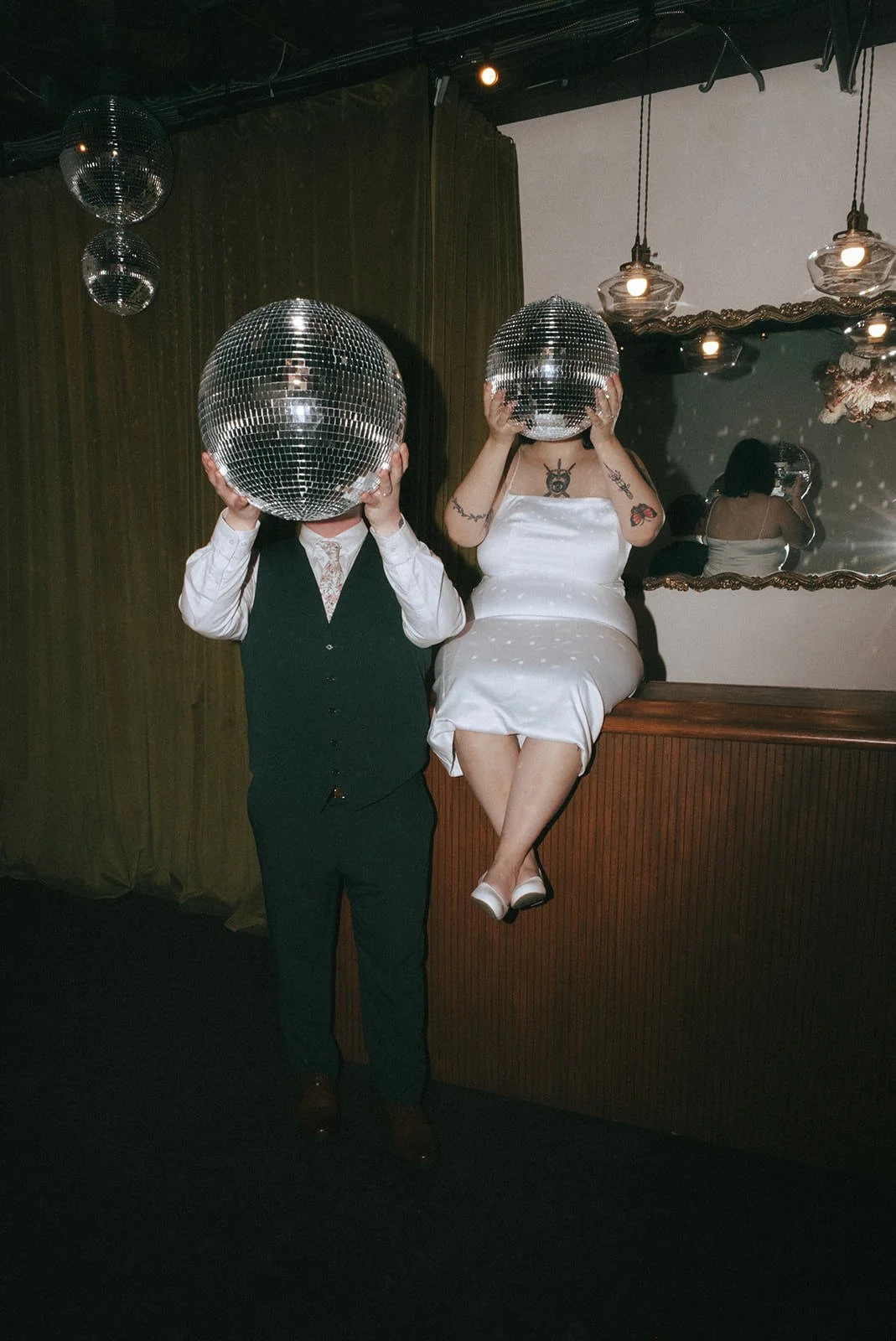 A newlywed sitting on a cabinet while their partner stands next to them as they both hold disco balls over their faces