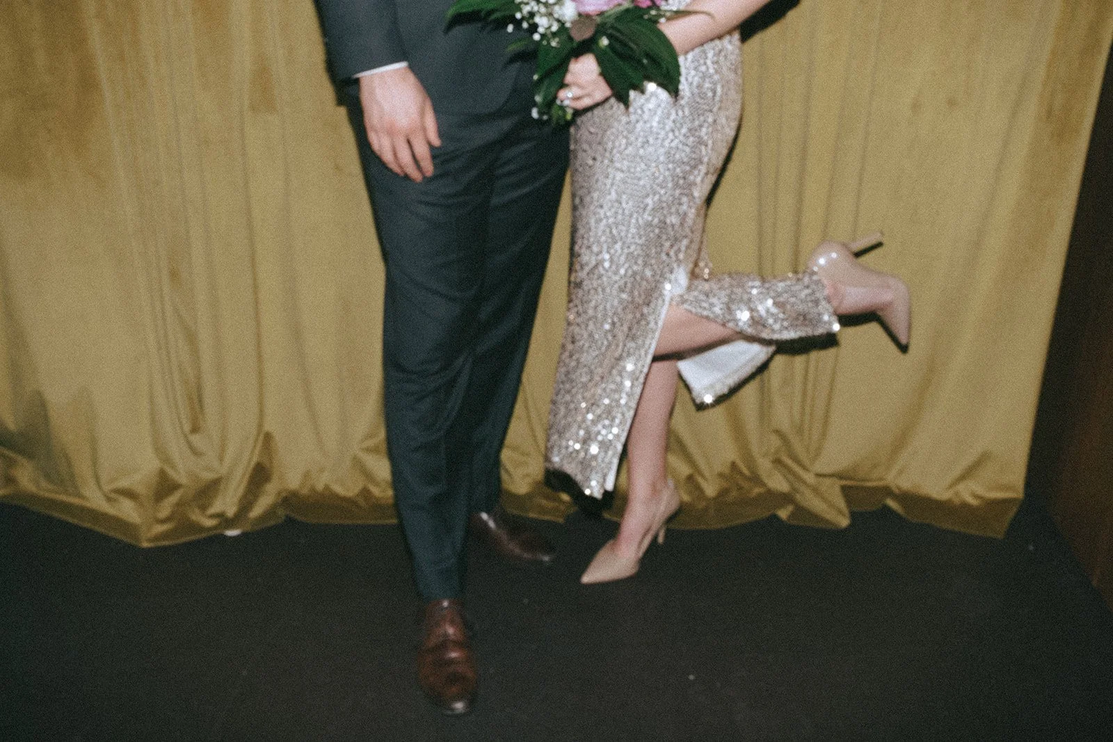 A close-up of a couple standing in front of a yellow curtain. One person is in a dark suit, and the another is in a sequined dress holding a bouquet. The second person is playfully lifting one leg, wearing nude high heels.